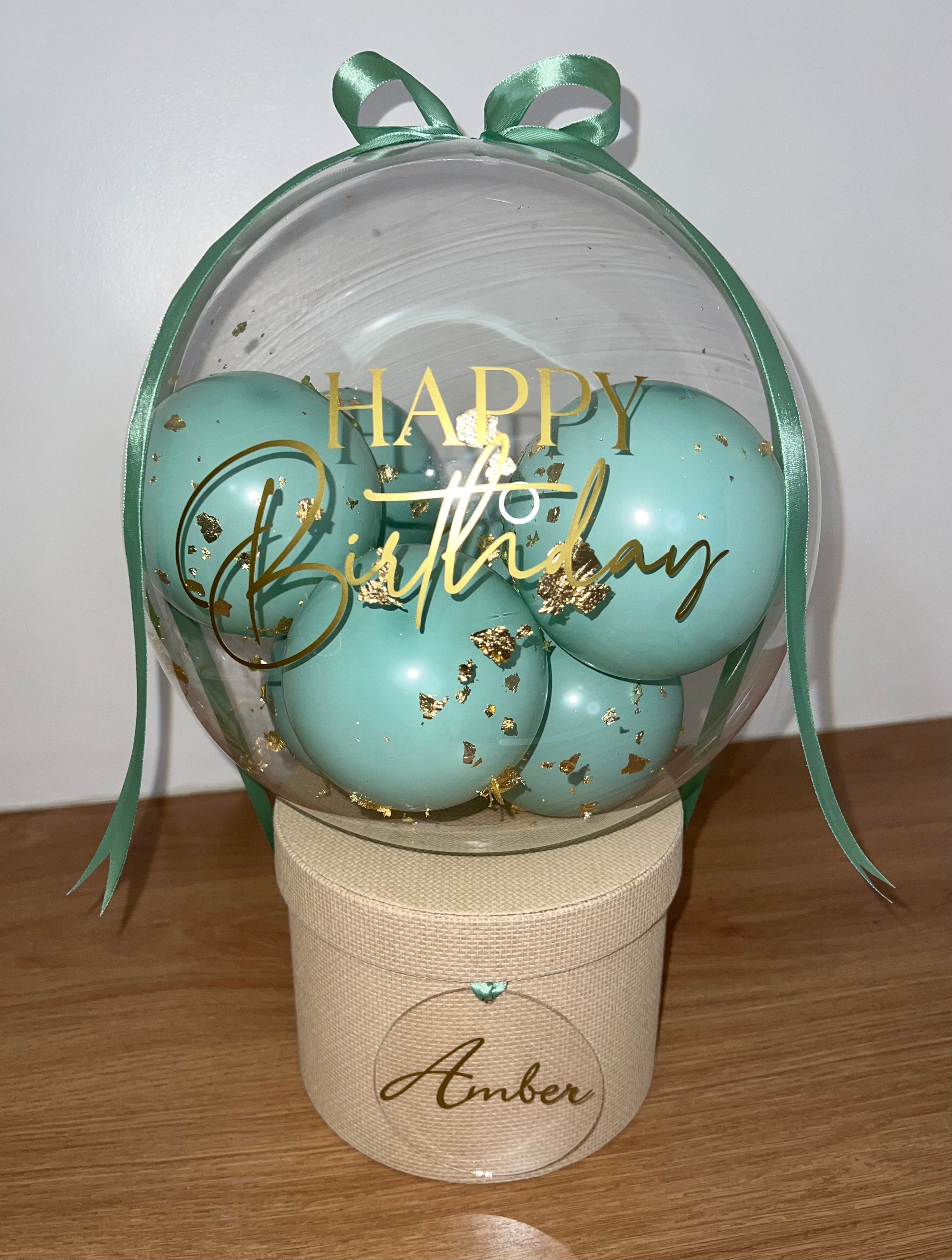 Beige textured hat box, with a clear 16inch bubble balloon on top secured with a sage green satin ribbon. Inside the clear balloon are multiple mini sage green balloons, with added gold flakes. The outside of the clear balloon ready happy birthday in elegant font with a matching acrylic disk attached to the hat box with the birthday girls name on (amber)