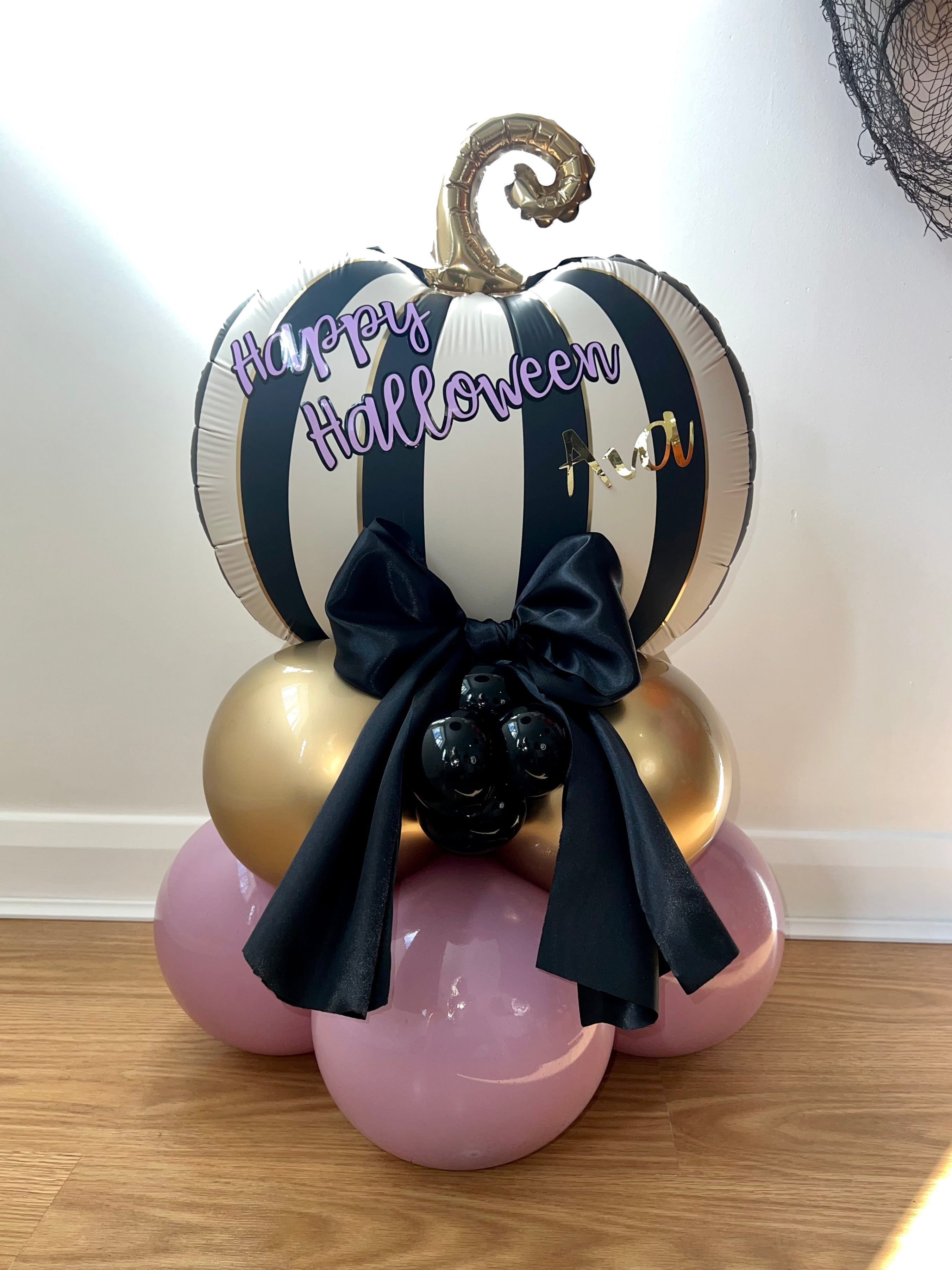 Halloween-themed balloon Stack decor with striped Pumpkin top, black bow, and Purple and gold balloons, with purple black and gold text saying Happy Halloween 'Ava'.