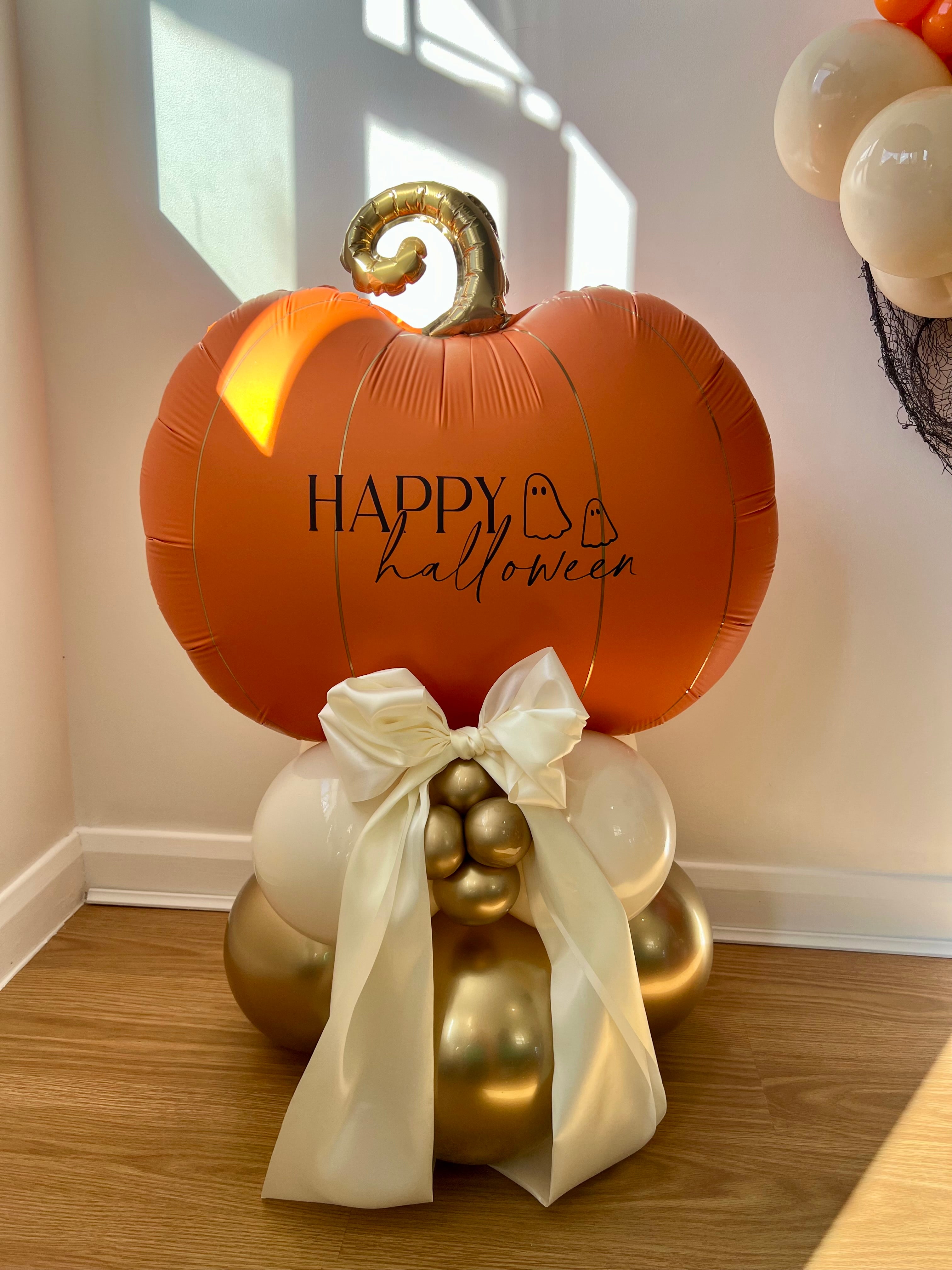 Decorative Halloween Balloon Stack arrangement with a pumpkin balloon, gold and white balloons on the base, and a white satin bow. Happy Halloween text with ghosts.